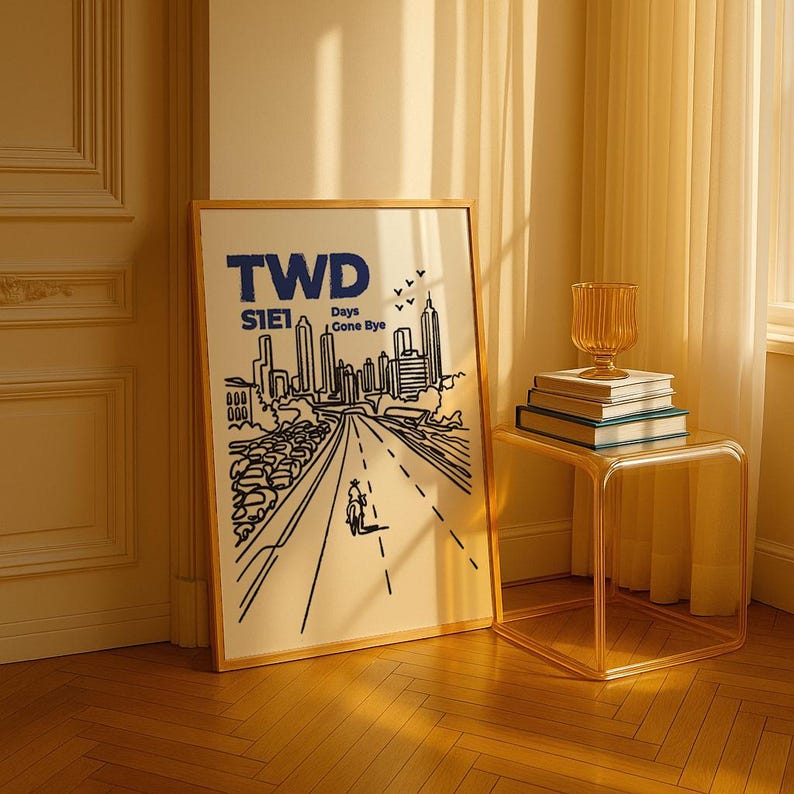 May include: A framed art print with the text "TWD S1E1 Days Gone Bye" in blue. The artwork depicts a road leading to a city skyline, with a figure on a motorcycle. A glass side table holds books and a decorative vase.