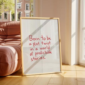 May include: A framed print with the quote "Born to be a plot twist in a world of predictable stories" in red lettering. The frame is gold, and the print is leaning against a wall near a window and a pink sofa.
