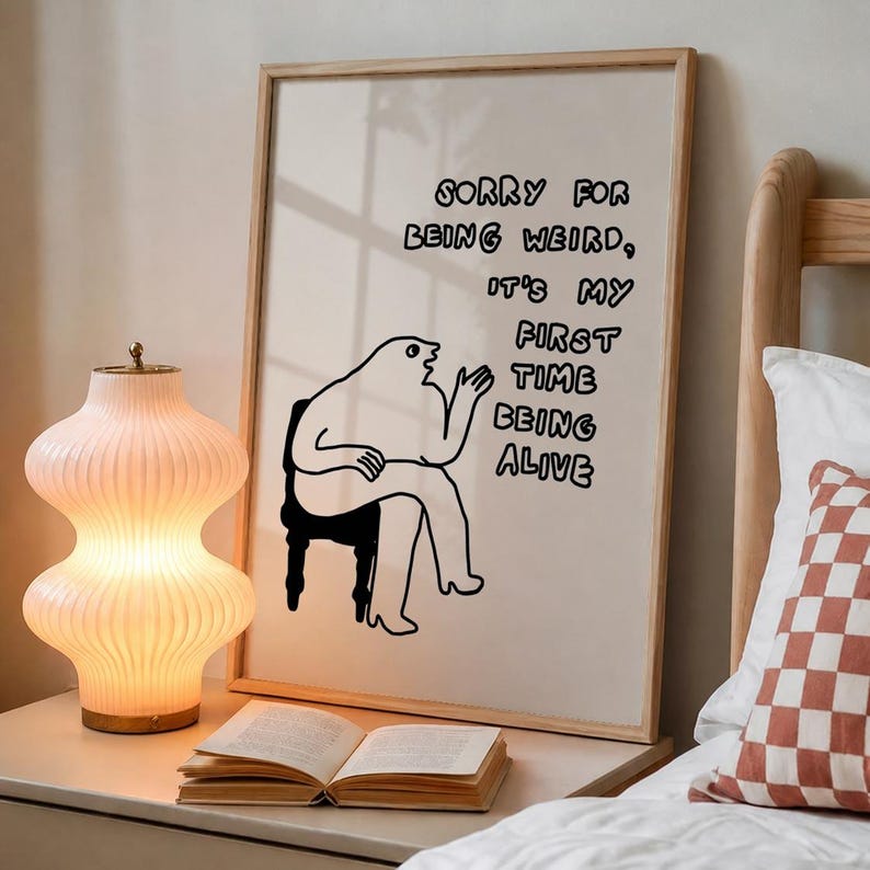 May include: A framed art print with the text "SORRY FOR BEING WEIRD, IT'S MY FIRST TIME BEING ALIVE" in black. The print features a cartoon figure sitting on a chair. A decorative lamp and an open book are on a bedside table.