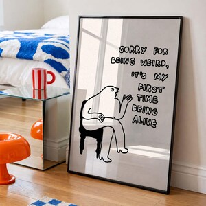 May include: A black framed print with a minimalist illustration of a person sitting on a chair. The text on the print reads: "SORRY FOR BEING WEIRD, IT'S MY FIRST TIME BEING ALIVE." The print is leaning against a wall.