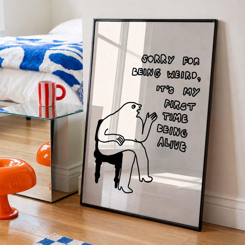 May include: A black framed print with a minimalist illustration of a person sitting on a chair. The text on the print reads: "SORRY FOR BEING WEIRD, IT'S MY FIRST TIME BEING ALIVE." The print is leaning against a wall.
