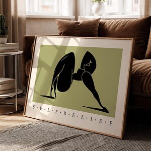 May include: A rectangular framed wall art print featuring a sage green background with a stylized black silhouette of a figure in a yoga pose. The text S E L F B E L I E F is printed in black capital letters along the bottom edge of the print.