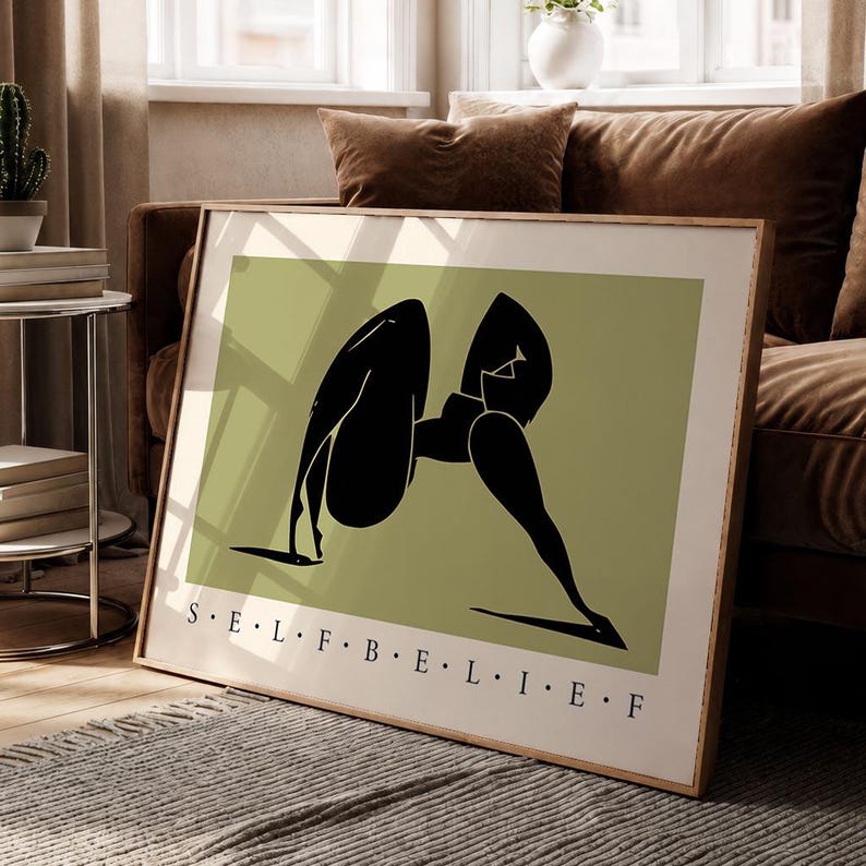 May include: A rectangular framed wall art print featuring a sage green background with a stylized black silhouette of a figure in a yoga pose. The text S E L F B E L I E F is printed in black capital letters along the bottom edge of the print.
