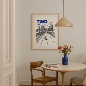 May include: A framed art print with a minimalist black and white cityscape illustration. The print features the text "TWD S1E1 Days Gone Bye" in blue. A table with a vase of flowers and a pendant light are in the foreground.