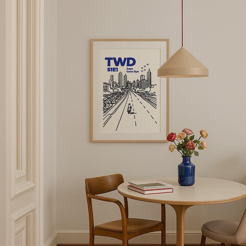 May include: A framed art print with a minimalist black and white cityscape illustration. The print features the text "TWD S1E1 Days Gone Bye" in blue. A table with a vase of flowers and a pendant light are in the foreground.