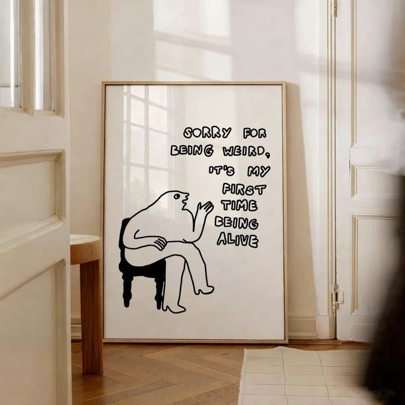 May include: A framed art print with a minimalist illustration of a seated figure. The black and white artwork features the text "SORRY FOR BEING WEIRD, IT'S MY FIRST TIME BEING ALIVE."