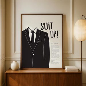 May include: A framed monochrome print displaying a suit illustration with the words "SUIT UP!". The artwork features a black suit jacket, tie, and shirt collar. The print is a stylish piece of wall art.