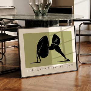 May include: A framed rectangular wall art print featuring a sage green background with a black abstract silhouette of a figure in a yoga pose. The text S E L F B E L I E F is printed in black capital letters at the bottom of the white border.