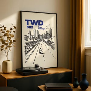 May include: Black framed art print with a minimalist illustration of a city skyline and a road with a person on a motorcycle. The text "TWD S1E1 Days Gone Bye" is in blue. A black record player sits below the art.
