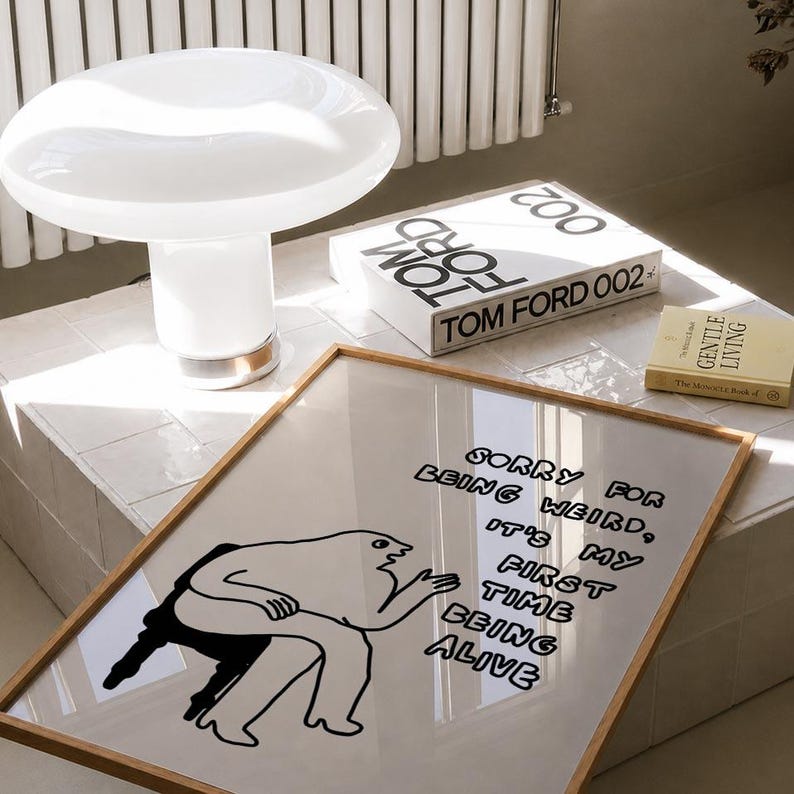 May include: A white mushroom-shaped table lamp with a gold base sits next to a framed art print. The print features a black and white illustration with the text "SORRY FOR BEING WEIRD, IT'S MY FIRST TIME BEING ALIVE". A Tom Ford book and a small book are also visible.