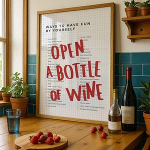 May include: A framed print with the text "OPEN A BOTTLE OF WINE" in red, with a list of activities. A bottle of red wine and a bottle of clear liquid are on a wooden counter, along with strawberries.
