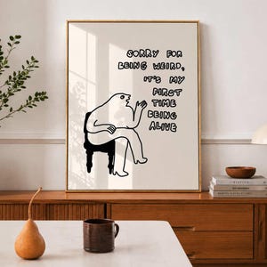 May include: A framed art print with a gold frame. The artwork features a whimsical illustration of a seated figure with the text "SORRY FOR BEING WEIRD, IT'S MY FIRST TIME BEING ALIVE". The figure is drawn in black and white.