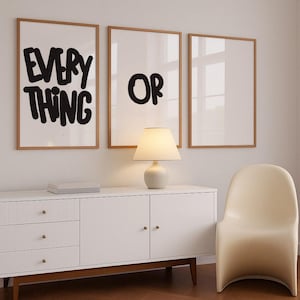 May include: Three framed wall art prints with wooden frames. The first print says "EVERY THING" in bold black letters. The second print says "OR" in black. A white lamp and a white cabinet are below the prints.