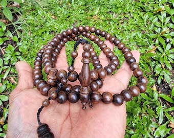 May include: A string of dark brown wooden prayer beads featuring a central carved wooden pendant and a smaller decorative bead at the end, all connected by a thin black cord, arranged in a circular pattern with smooth, round beads.