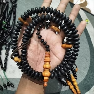 Peut inclure: Un ensemble de chapelets noirs avec des perles ovales et des accents en bois. Les perles sont enfilées avec un cordon foncé et comportent des séparateurs en bois décoratifs et un pendentif en forme de gland. Les perles sont tenues dans une main.