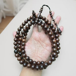 May include: A dark brown wooden prayer beads, also known as a misbaha or tasbih, with multiple strands of round beads. The beads are held in a hand, showcasing the natural wood grain and texture. The beads are connected by a dark string.