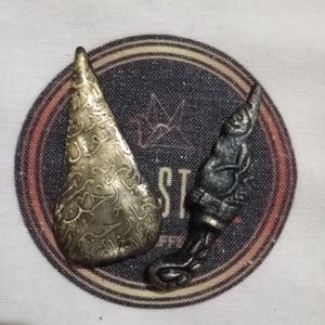 Könnte beinhalten: Zwei Metallskulpturen auf dunklem Hintergrund. Eine ist ein goldfarbenes, tropfenförmiges Stück mit aufwendigen eingravierten Mustern. Die andere ist eine dunkelgraue, stilisierte Figur mit detaillierten Schnitzereien, möglicherweise eine kleine Statue oder ein Anhänger.