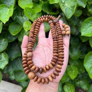 Puede incluir: Un collar de cuentas de oración de madera con un acabado marrón natural. El collar presenta múltiples hilos de cuentas redondeadas, con cuentas más grandes y prominentes intercaladas. Las cuentas se sostienen en una mano contra un fondo de hojas verdes.