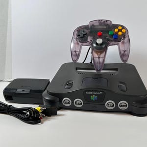 Peut inclure: Ensemble complet de console de jeu Nintendo 64. Comprend une console grise, une manette violette translucide, un adaptateur secteur noir et un câble AV noir. La console porte le logo Nintendo 64.