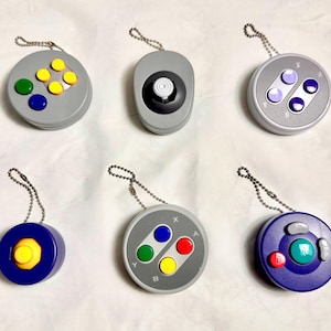 Könnte beinhalten: Sechs Schlüsselanhänger mit Miniatur-Gamecontrollern. Die Controller haben verschiedene Tastenanordnungen und Farben, darunter Grau, Blau, Gelb, Grün und Rot. Jeder Schlüsselanhänger hat eine silberne Kette zur Befestigung.