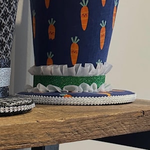 May include: A navy blue top hat with a carrot pattern, white lace trim, and a green glitter band. The hat has a white ruffled trim and is sitting on a wooden shelf. A similar hat is visible below.