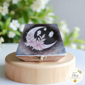 Peut inclure: Une boîte à bijoux en bois avec un motif céleste. Le couvercle présente un croissant de lune, des cristaux et des fleurs roses sur un fond sombre. La boîte est présentée sur un socle en bois et possède un fermoir en métal.