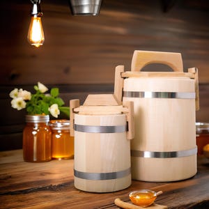 Puede incluir: Dos recipientes de madera para almacenar miel con bandas metálicas, uno grande y otro pequeño, sobre una superficie de madera. También hay tarros de miel y un cuenco de panal, lo que sugiere un tema de apicultura. Un pequeño cuenco de miel y una cuchara de madera en primer plano.