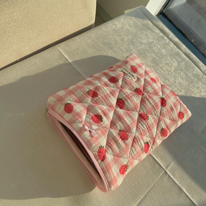 May include: Pink quilted laptop sleeve with a white and pink gingham pattern and a strawberry print. The sleeve has a zipper closure and the text "TWINLOOK" is printed on the front.