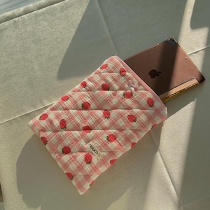 Peut inclure: Une housse de tablette matelassée rose et blanche avec un imprimé de fraises et le texte "mimuco" sur le devant. La housse est posée sur une surface blanche avec une tablette à l'intérieur.