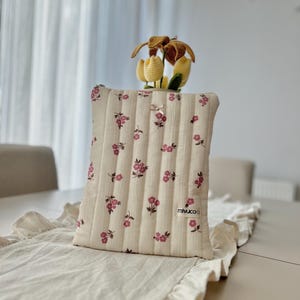 May include: A cream-coloured, quilted pillow with a floral pattern of pink flowers. The pillow is rectangular and has vertical stitching. A small, crocheted flower arrangement sits atop the pillow. The pillow is on a cream-coloured table runner.