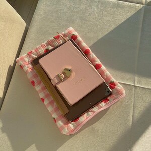 May include: A pink and white quilted fabric pouch with a strawberry pattern. The pouch contains a pink journal with a gold lock and the words "little diary" on the cover, a brown notebook, and a tablet.