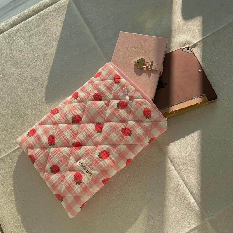 May include: A pink and white quilted pouch with a strawberry print and the word "minuco" on it. The pouch is open and has a pink notebook and a pink tablet inside.