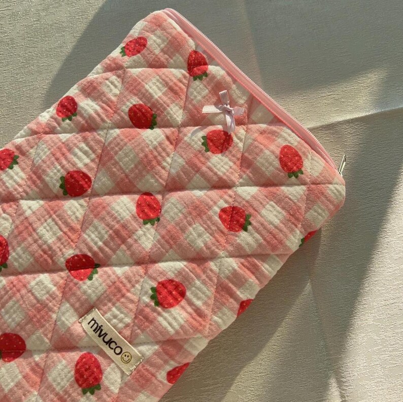 May include: Pink quilted zippered pouch with a white and pink gingham pattern and embroidered strawberries. The pouch has a pink ribbon bow and a white label with the text "miwuco" and a smiley face.
