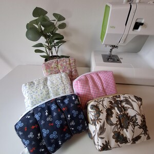 Pode incluir: Uma coleção de bolsas de tecido feitas à mão em vários padrões e cores. Os designs incluem motivos florais, xadrez e celtas. Uma máquina de costura é visível no fundo, sugerindo um tema artesanal ou de bricolage.