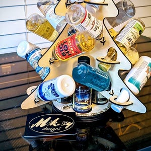May include: A wooden tree-shaped bottle display stand holding various liquor bottles. The stand is made of light-colored wood and features multiple tiers to hold the bottles. The base has a black label with the text "Mik.84 designs".
