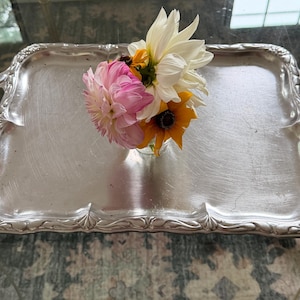 May include: A rectangular silver-toned metal serving tray with ornate, embossed floral handles and edges. A small glass vase sits in the center of the tray, holding a bouquet of fresh flowers including white, pink, and yellow dahlia-like blooms.