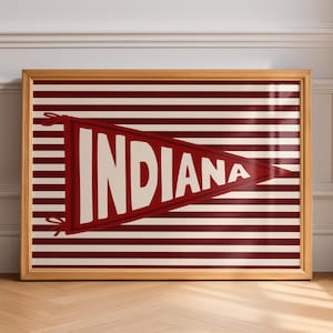 May include: A framed horizontal print featuring a dark red triangular pennant with the word INDIANA in large white capital letters. The pennant is centered over a background of alternating horizontal dark red and white stripes.
