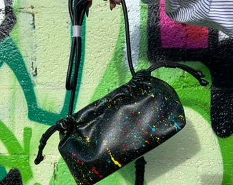 Puede incluir: Bolso bandolera negro con diseño de salpicaduras de pintura coloridas. El bolso tiene un cierre de cordón y una larga correa negra. El fondo es una pared cubierta de graffiti con pintura verde, rosa y negra.