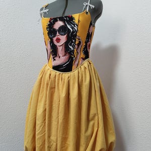 May include: A yellow dress with a fitted bodice featuring a pop art style illustration of a woman wearing sunglasses. The dress has a gathered, voluminous skirt. The straps are white with small bows. The dress is displayed on a mannequin.