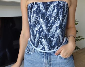 May include: A strapless corset top with a dark blue and white bird pattern. The corset is paired with light blue denim jeans. The top has a white trim along the top and bottom edges.