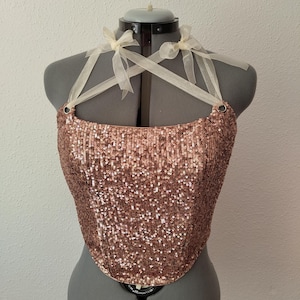Può includere: Un top a corsetto con paillettes color rame esposto su un manichino. Il top presenta una scollatura a cuore ed è adornato da piccole paillettes scintillanti. Delicati nastri trasparenti sono legati alle spalle, aggiungendo un tocco di eleganza.