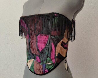 May include: A colorful corset with a black fringe trim. The corset features a vibrant pattern of pink, green, and brown shapes on a dark background. The garment is displayed on a gray dress form.