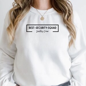 Puede incluir: Sudadera blanca de cuello redondo con el texto "BEST.SECURITY.SQUAD" dentro de un rectángulo negro, con "Smith's Crew" debajo. La sudadera es de color blanco liso.