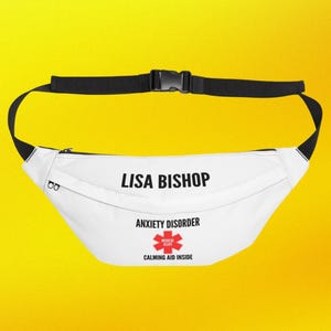 Könnte beinhalten: Weiße Bauchtasche mit verstellbarem schwarzem Riemen und Schnalle. Auf der Vorderseite ist der Text "LISA BISHOP" aufgedruckt, zusammen mit "ANXIETY DISORDER" und einem medizinischen Warnsymbol. Die Worte "CALMING AID INSIDE" sind ebenfalls auf der Vorderseite aufgedruckt.