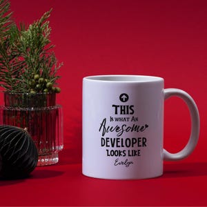 Op de afbeelding: Witte keramische mok met de tekst "THIS is WHAT AN Awesome DEVELOPER LOOKS LIKE Evelyn" in zwart schrift. De mok heeft een oor en staat tegen een rode achtergrond.