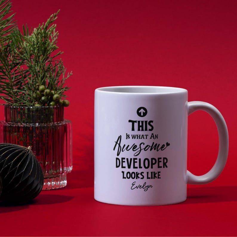 Op de afbeelding: Witte keramische mok met de tekst "THIS is WHAT AN Awesome DEVELOPER LOOKS LIKE Evelyn" in zwart schrift. De mok heeft een oor en staat tegen een rode achtergrond.