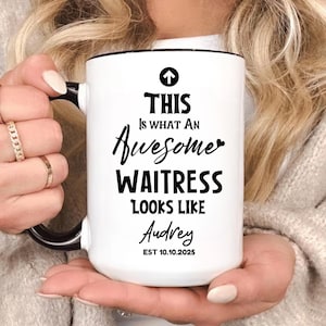 May include: White ceramic mug with a black handle and rim. The mug features the text "THIS Is WHAT AN Awesome WAITRESS LOOKS LIKE Audrey EST 10.10.2025" in black script and bold fonts. An arrow points upwards.