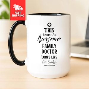 Puede incluir: Taza de cerámica blanca con asa y borde negros. La taza presenta el texto "THIS IS WHAT AN Awesome FAMILY DOCTOR LOOKS LIKE Dr. Evelyn EST 10.10.2025" en escritura negra. Un rectángulo rojo con las palabras "FAST SHIPPING" está en la esquina superior izquierda.