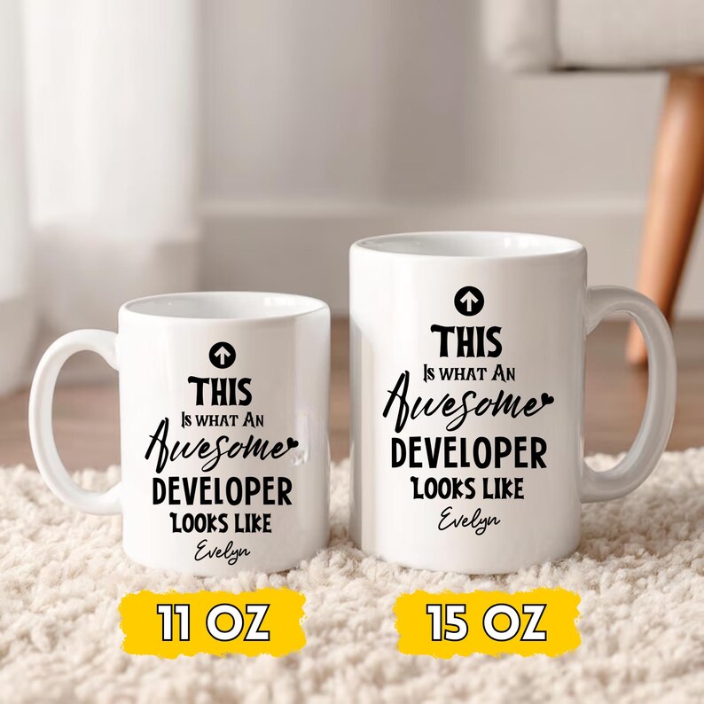 Op de afbeelding: Twee witte keramische mokken met zwarte tekst: "THIS IS WHAT AN Awesome DEVELOPER LOOKS LIKE Evelyn." E&eacute;n mok is 325 ml, de andere is 444 ml. Beide hebben een zwarte pijl boven de tekst.