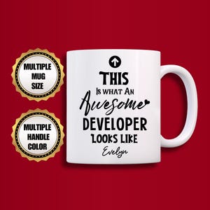 Op de afbeelding: Witte keramische mok met een zwarte pijl en de tekst "THIS IS WHAT AN Awesome DEVELOPER LOOKS LIKE Evelyn". De mok heeft een wit handvat en staat tegen een rode achtergrond. Twee goudkleurige cirkelvormige badges staan links.